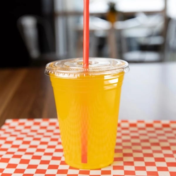 A bright yellow drink with a red straw in a clear plastic cup on a red and white checkered paper.
