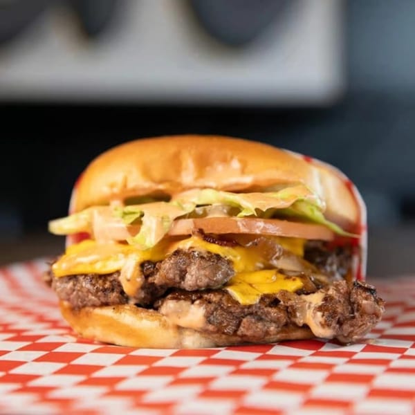 A SMA5H double cheeseburger with lettuce, tomato, and sauce on a red and white checkered wrapper.