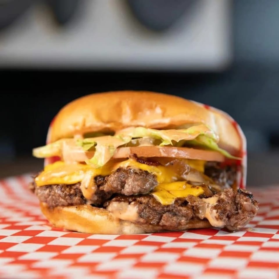 A SMA5H double cheeseburger with lettuce, tomato, and sauce on a red and white checkered wrapper.