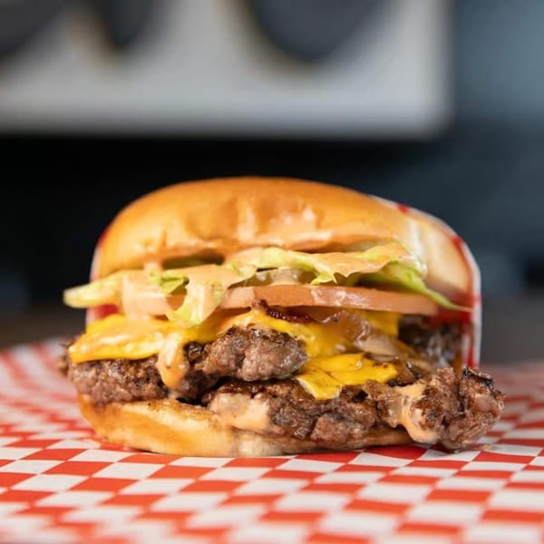 A SMA5H double smash burger with melted cheese, lettuce, tomato, and sauce on a red checkered paper.