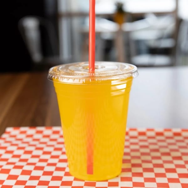 A bright yellow drink in a clear plastic cup with a red straw on a red and white checkered paper.