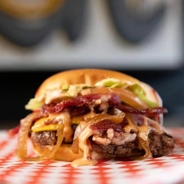 A SMA5H burger with bacon, caramelized onions, cheese, and sauce on a red and white checkered paper.